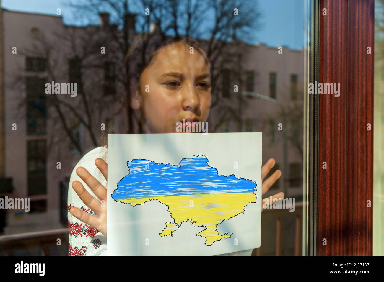 Una chica con un vestido bordado mira por la ventana, sostiene una sábana blanca con una foto y una inscripción. La niña protesta contra la guerra.Ukrainia Foto de stock