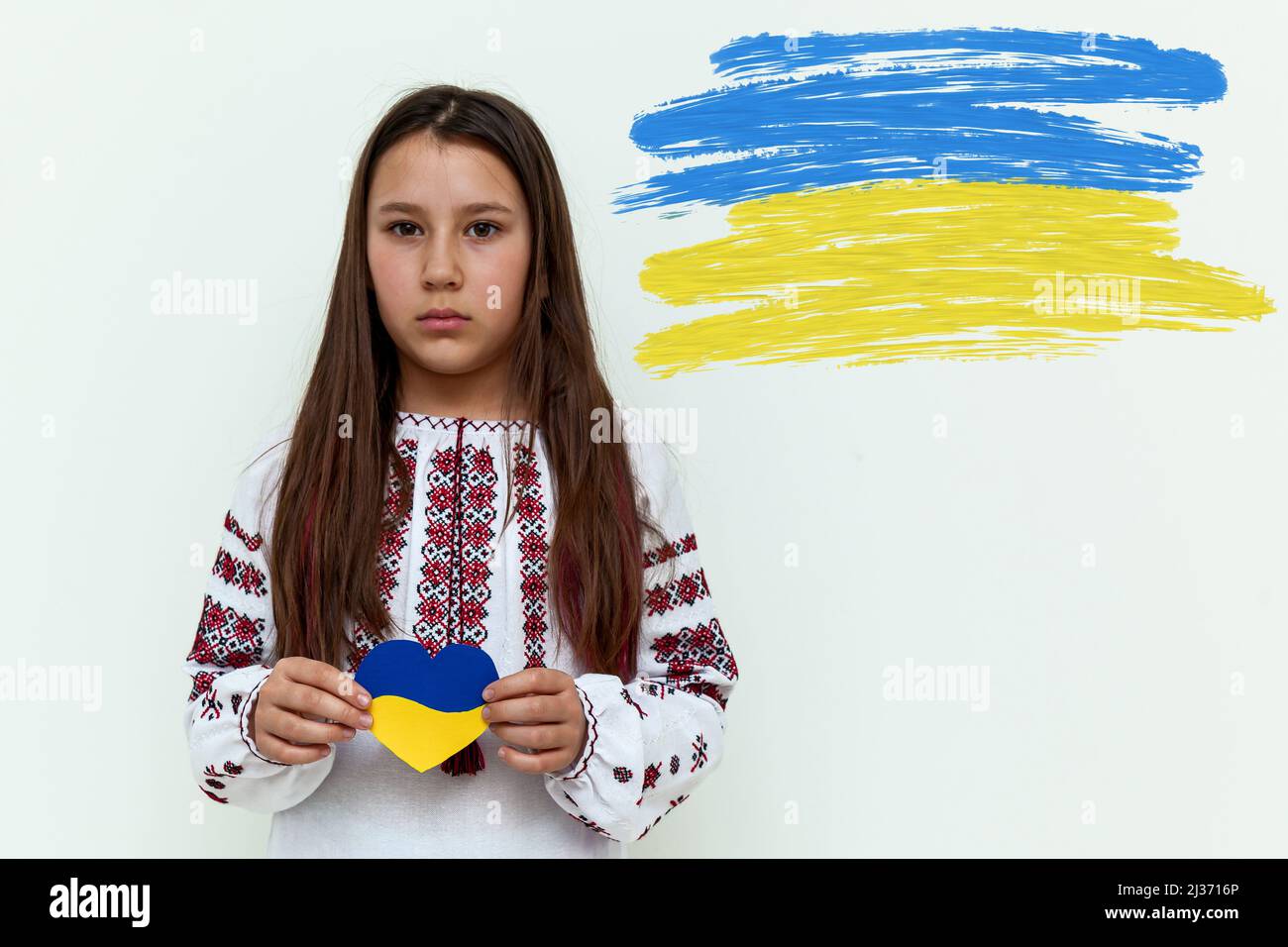 Una chica con un vestido bordado se levanta contra una pared blanca, sosteniendo un corazón pintado de azul y amarillo. La chica protesta contra la guerra y muestra lo Foto de stock