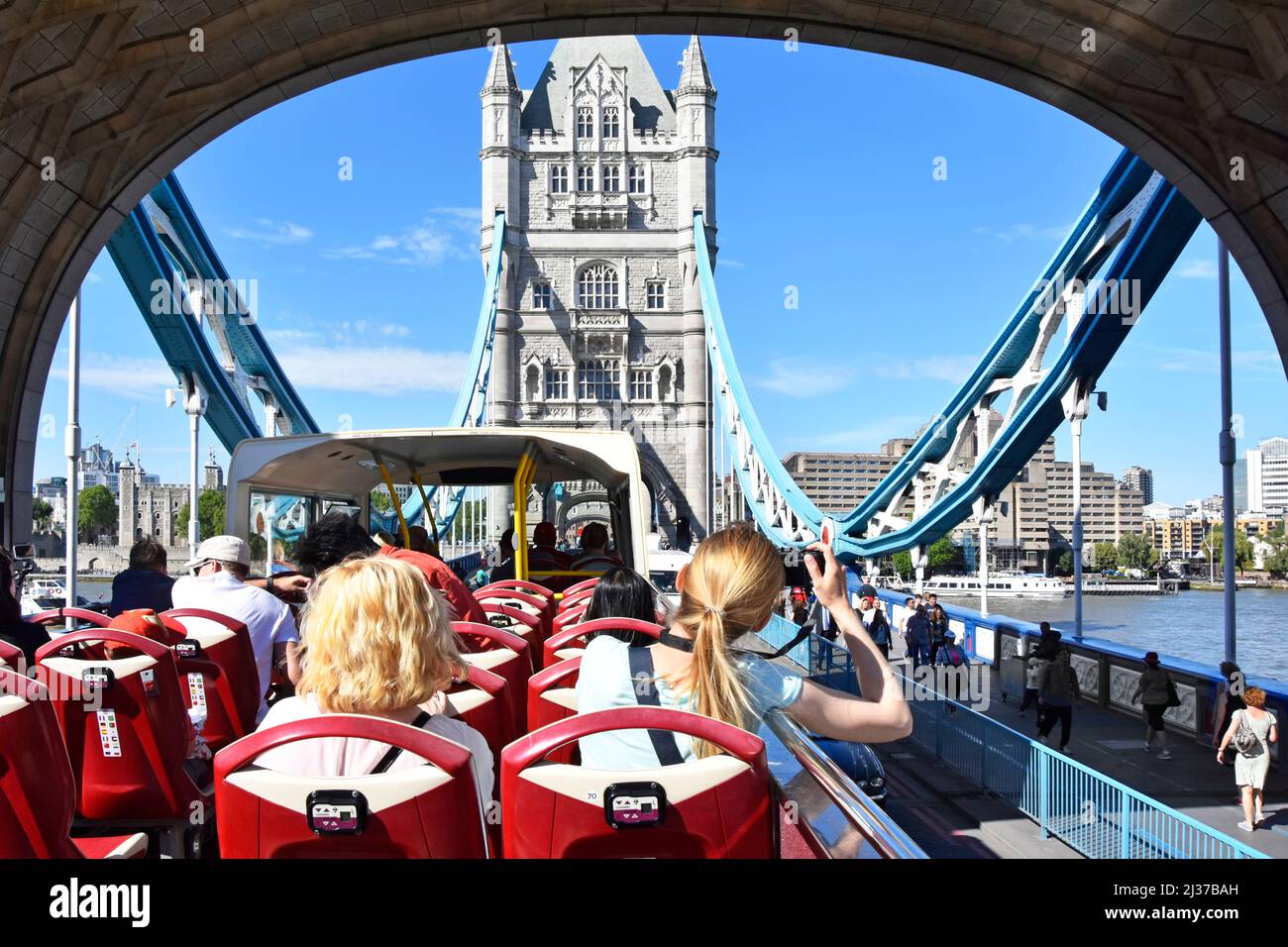 Volver ver a las mujeres en autobús turístico de dos pisos descubierto Londres tour bajo el arco de la histórica carretera Tower Bridge cruzando la marea alta río Támesis Inglaterra Reino Unido Foto de stock