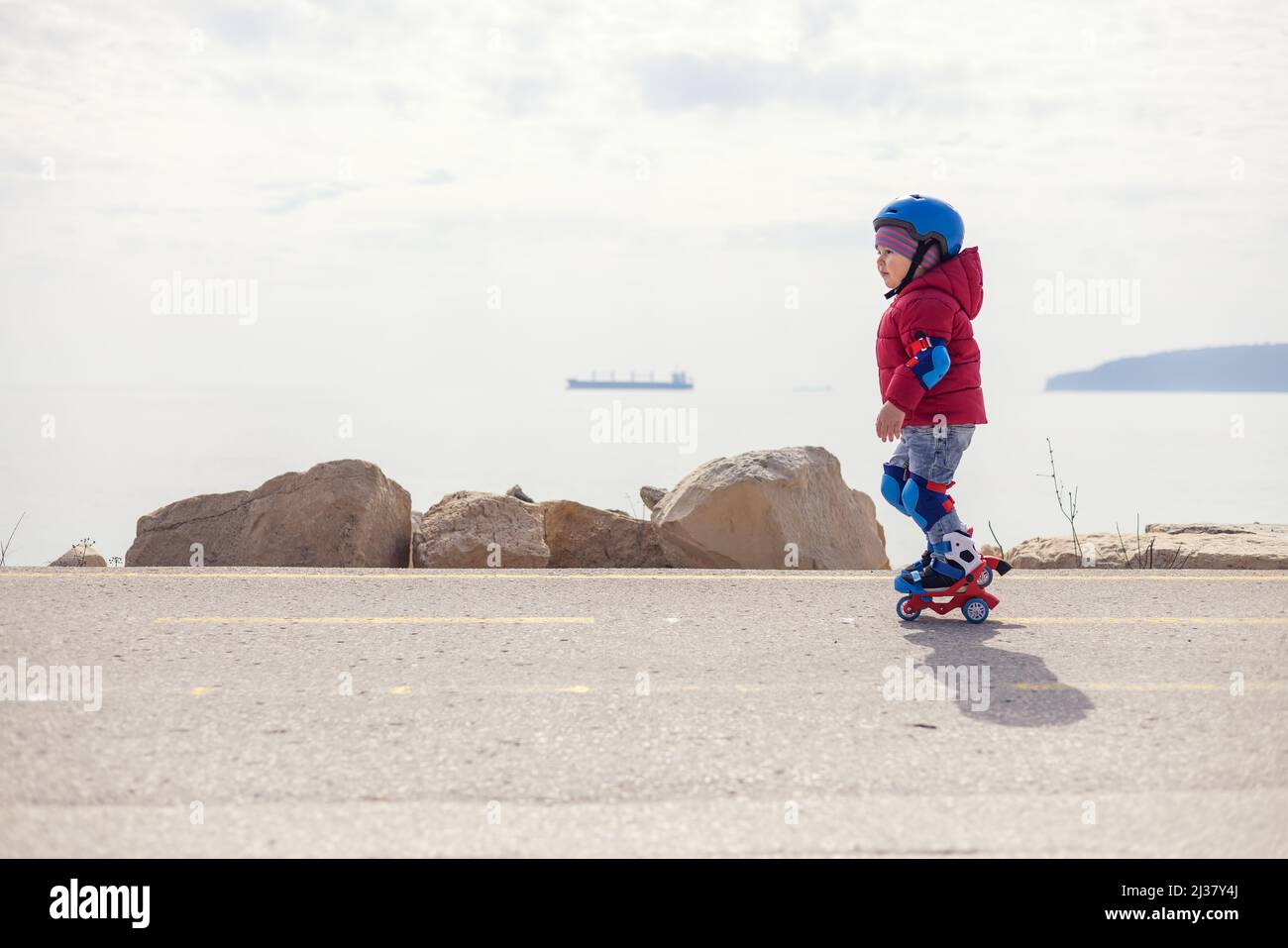 Niño pequeño que monta patines cerca de la orilla del mar. Niño con casco y protección en las cuchillas de rodillo. Patinaje sobre ruedas. Foto de stock