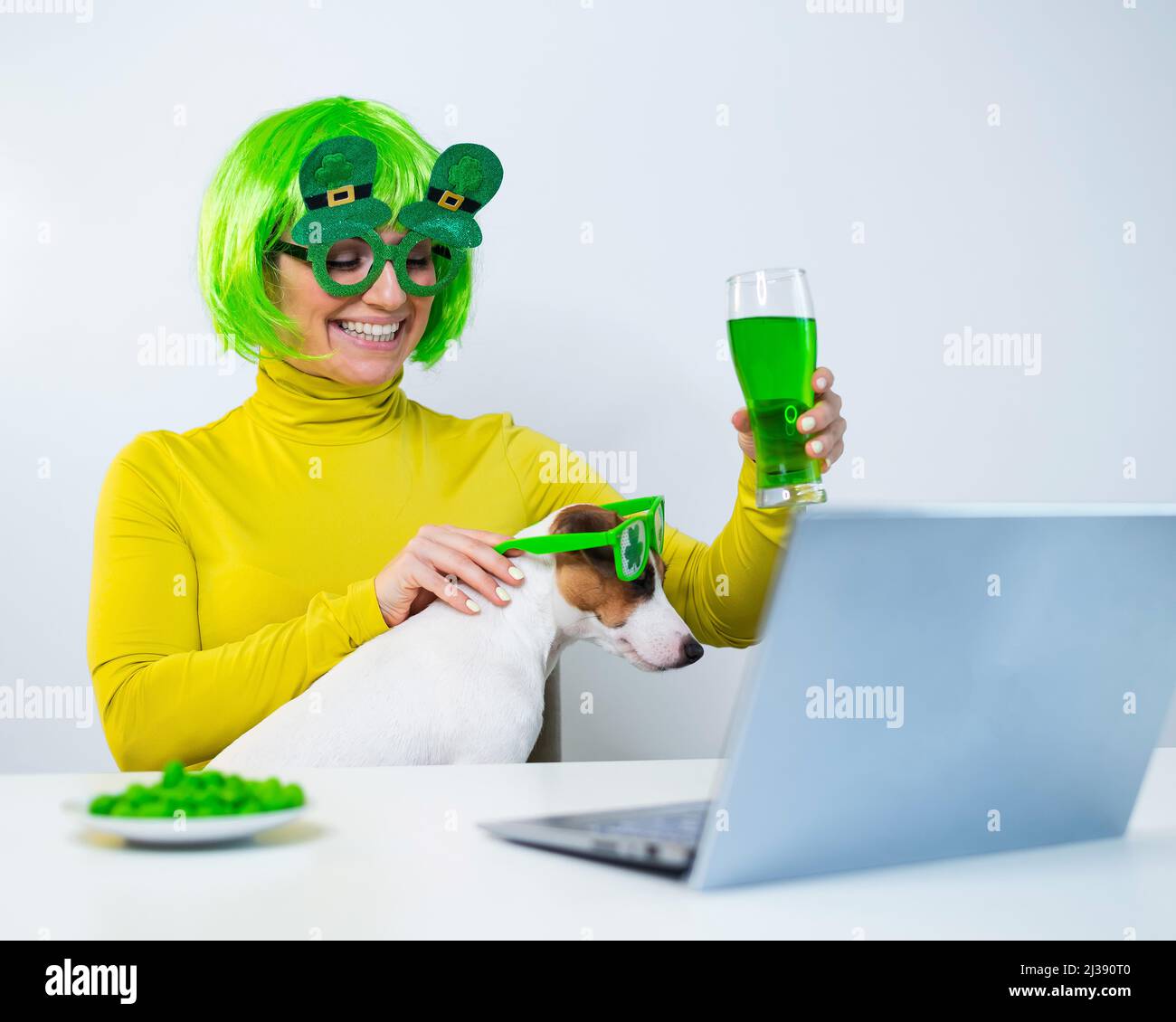 Una mujer joven en una peluca verde y vasos alegres bebe cerveza y pica nueces glaseadas. Una chica se sienta con un perro en una mesa y celebra el día de San patricio Foto de stock