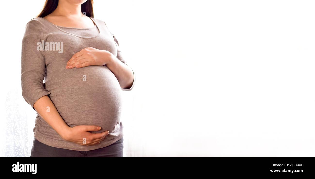 Mujer embarazada con vientre grande en la ventana. Concepto de atención prenatal de maternidad y embarazo de la mujer. Una mujer joven embarazada sostiene las manos sobre su vientre hinchado. Foto de stock