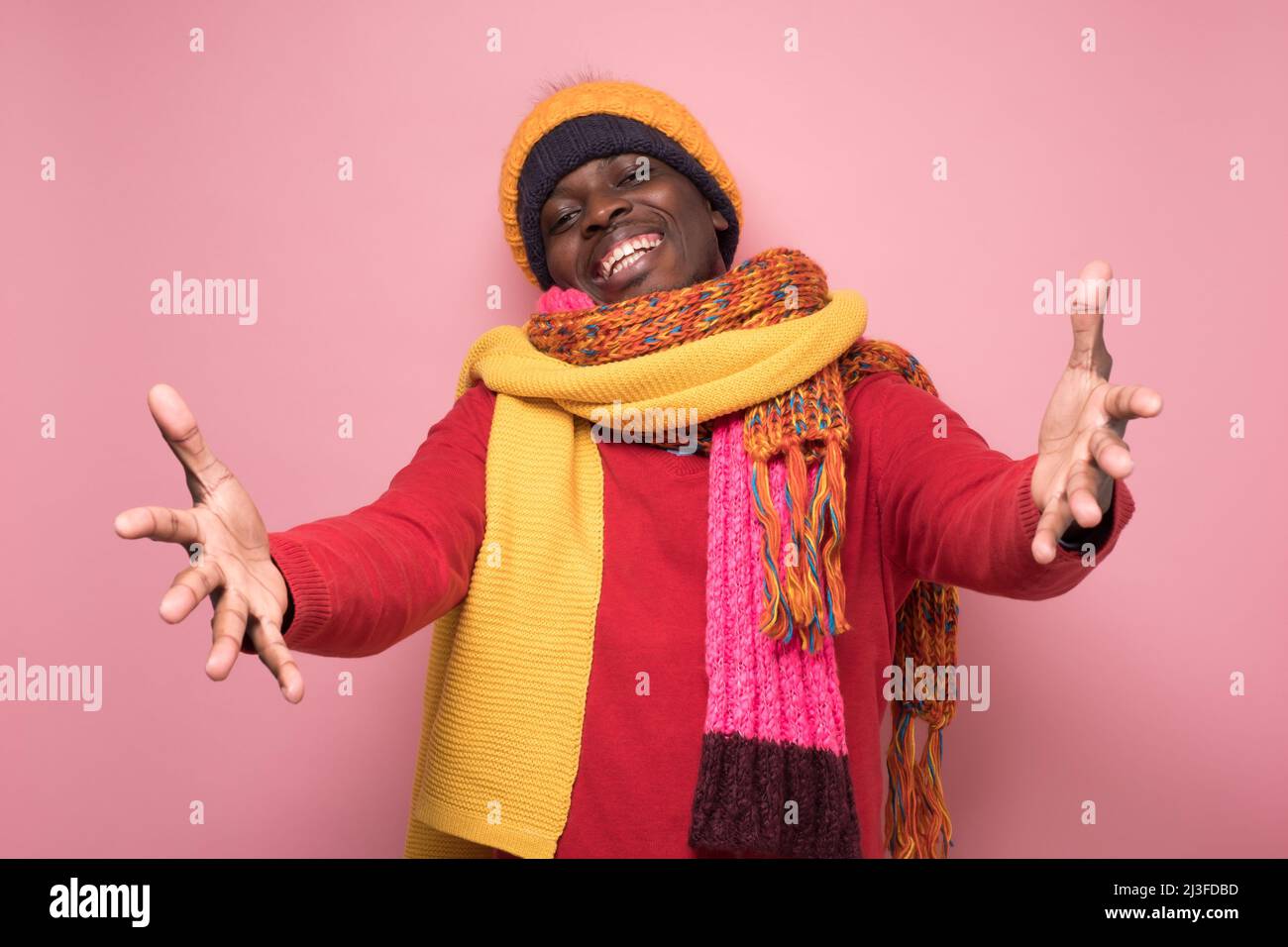 Hombre africano alegre vestido con varios sombreros y bufandas tirando de las manos hacia la cámara, queriendo acurrucarse, sonriendo ampliamente, amigo acogedor para entrar Foto de stock