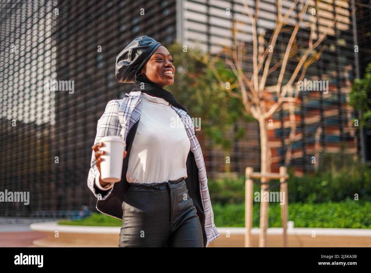 Mujer afroamericana positiva con ropa informal elegante y pañuelo tradicional caminando con bolso y café para llevar mientras se está apresurando a trabajar en el distrito moderno de la ciudad Foto de stock