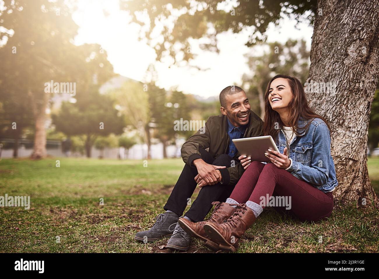 Mañanas tranquilas juntas. Foto de una alegre pareja joven sentada bajo un árbol con una tableta digital fuera en un parque. Foto de stock