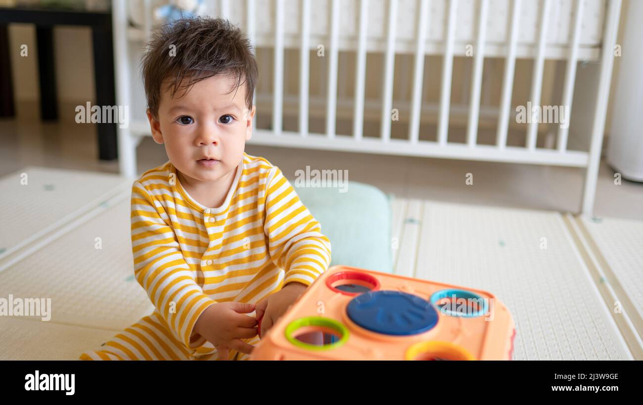 8 meses de edad raza mixta bebé niño jugando con caja de actividad en el dormitorio con pijama amarillo mientras se sienta en el suelo cubierto con un juego de goma Foto de stock