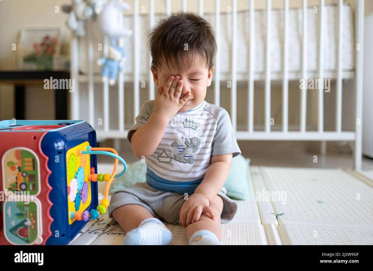 Soñoliento niño de 8 meses de edad de raza mixta jugando con una caja de actividades en el dormitorio mientras está sentado en el suelo cubierto con una alfombrilla de goma Foto de stock