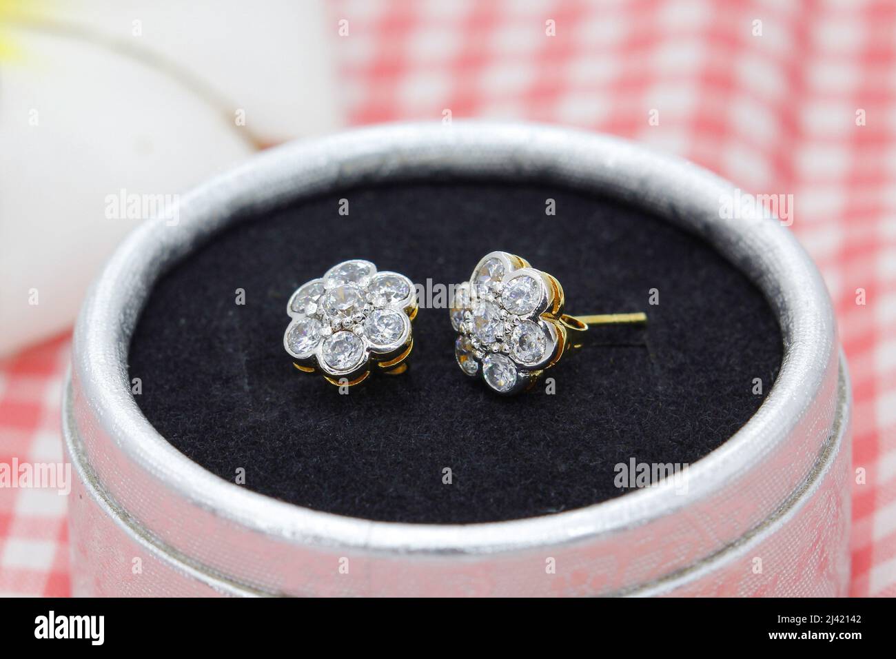 Hermosos pendientes dorados decorados con diamante en forma de flor se muestran en caja de joyas sobre fondo de tela de cuadros rojos Foto de stock