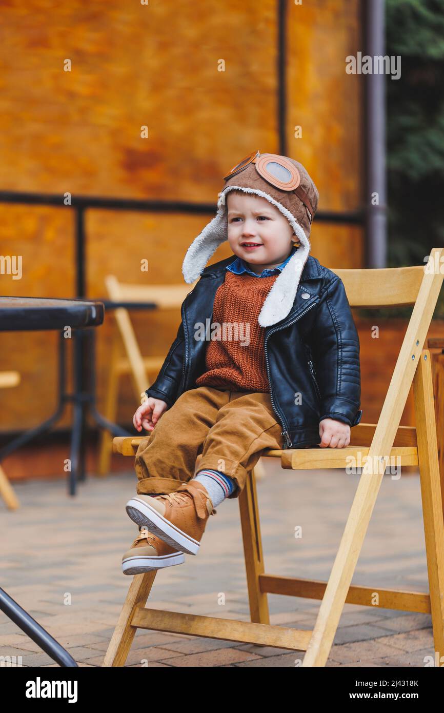 Niño de 3 años con con chaqueta de cuero y pantalón marrón sentado en la terraza cerca la cafetería. Niño moderno. Moda infantil. Niño feliz Fotografía de stock - Alamy