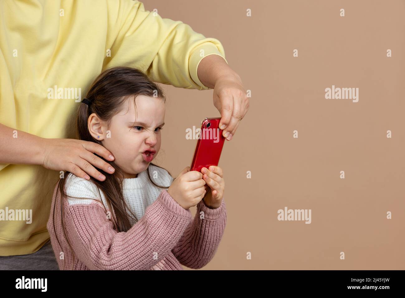 Retrato de los padres alejando el smartphone de las manos de las niñas, fondo beige. Hija enojada sostenga el teléfono apretado y resista. Concepto de prevención de Foto de stock