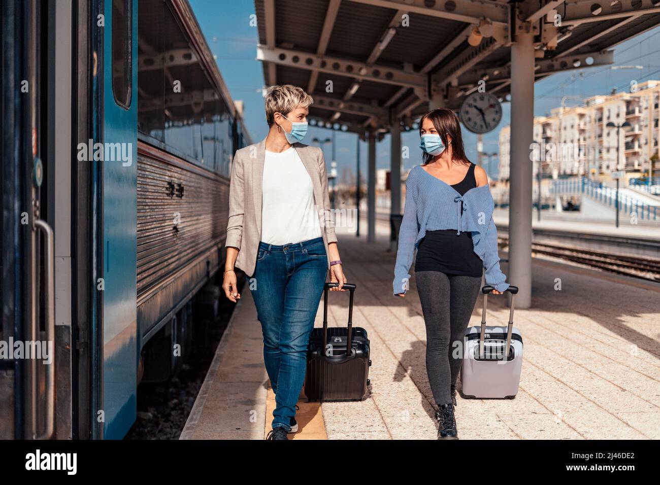 Dos felices amigas en máscaras protectoras caminando y charlando en la plataforma en la estación de tren Foto de stock