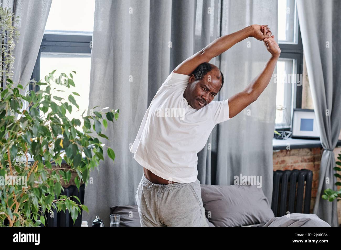 Hombre anciano contemporáneo en pijamas haciendo curvas laterales después de dormir mientras se encuentra junto a una cama doble en el dormitorio con cortinas grises Foto de stock