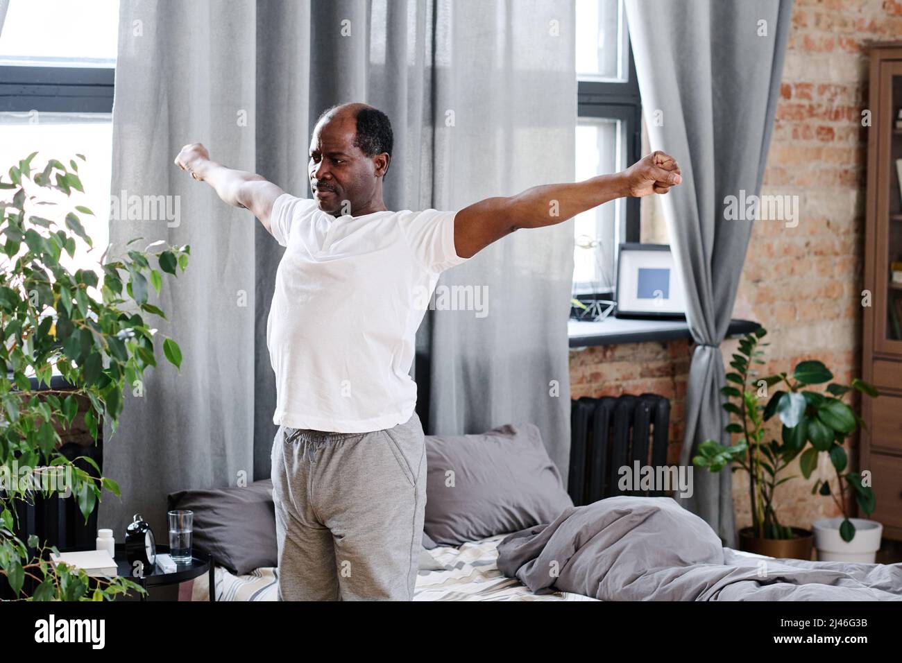 Hombre negro anciano activo con brazos extendidos haciendo ejercicio por la mañana mientras está de pie junto a su cama con almohadas grises y manta Foto de stock