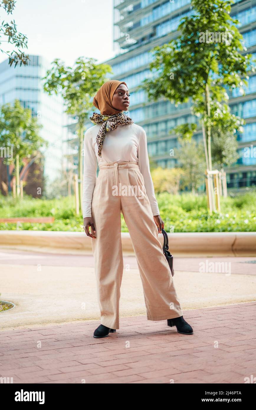 Cuerpo entero de mujer con seguro vestido con un elegante traje sport y un  tradicional hijab islámico con bufanda en el cuello de pie en el parque y  mirando hacia fuera Fotografía