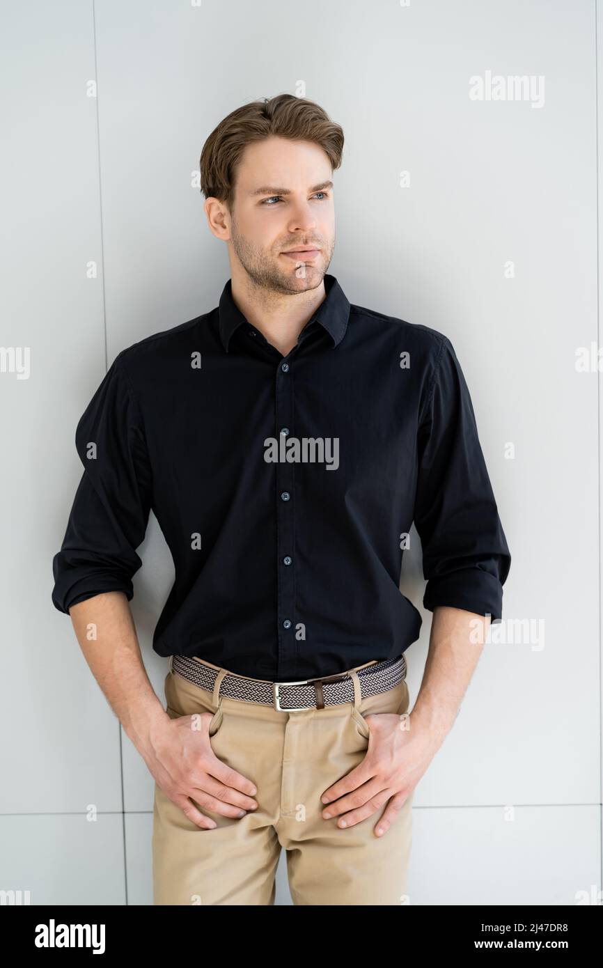 hombre con estilo con camisa negra y pantalón beige con pulgares en los  bolsillos cerca de la pared gris Fotografía de stock - Alamy