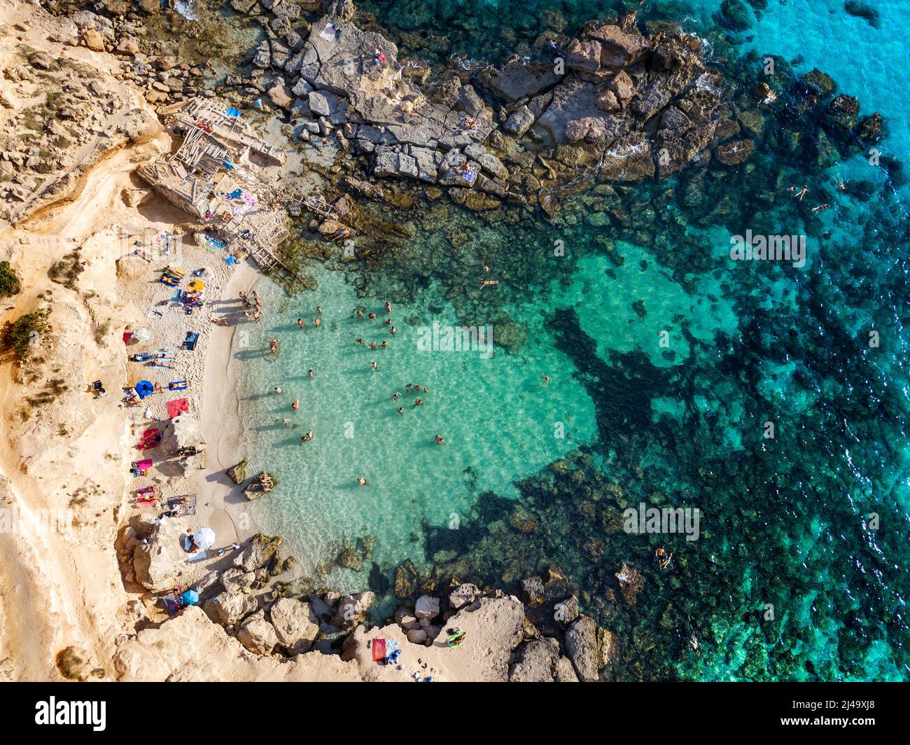 Playa de Es Caló d'es mort cala, vista aérea, visitantes veraniegos tumbados en una cala bordeada de acantilados rojos y rocas en un hueco de aguas turquesas, Formentera Ba Foto de stock