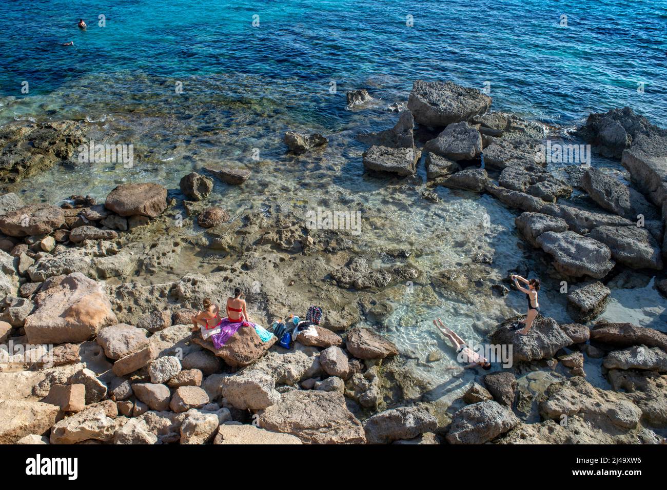Playa de Es Caló d'es mort cala, visitantes veraniegos tumbados en una cala bordeada de acantilados rojos y rocas en un hueco de aguas turquesas, isla de Formentera Baleares Foto de stock