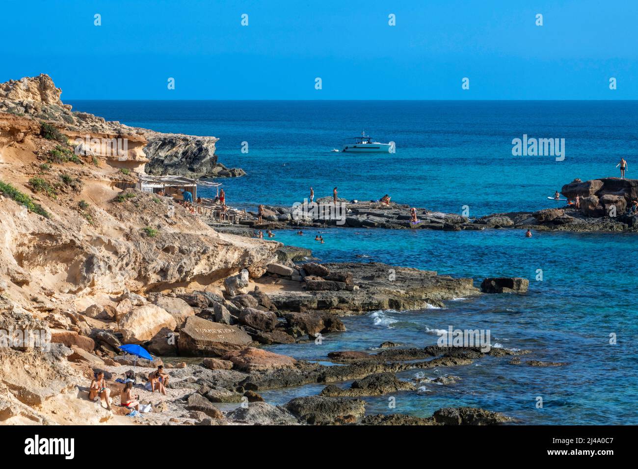Playa de Es Caló d'es mort cala, visitantes veraniegos tumbados en una cala bordeada de acantilados rojos y rocas en un hueco de aguas turquesas, isla de Formentera Baleares Foto de stock