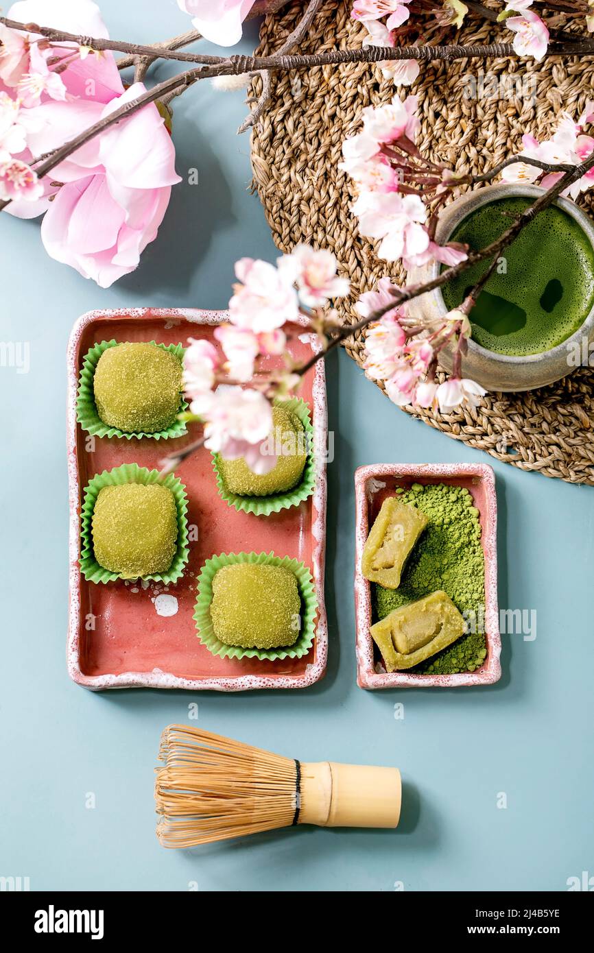 Postre de arroz tradicional asiático mochi de matcha verde dulce en plato de cerámica rosa con taza de té mate helado sobre mesa azul con flores rosadas de primavera. FL Foto de stock