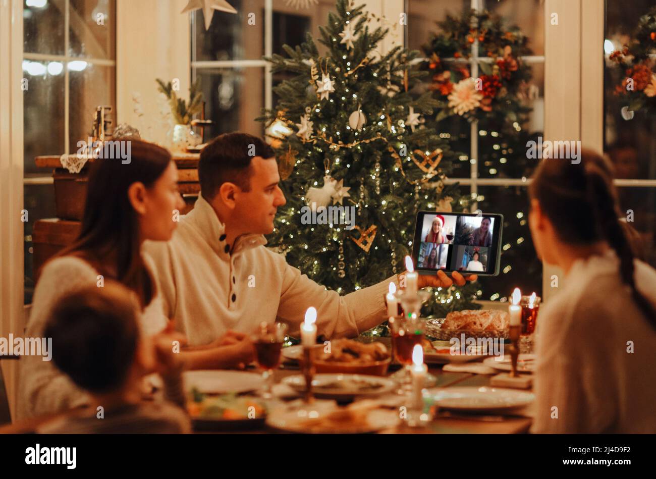 Feliz familia está sentado una mesa festiva en la sala de estar y teniendo una videollamada vía tablet con sus amigos. La habitación está decorada en el Cristo Foto de stock