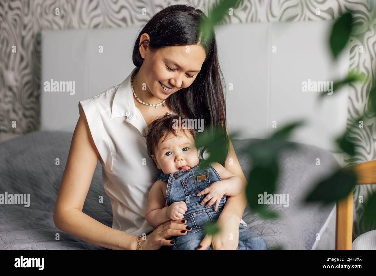 Retrato de madre feliz con hija pequeña en el dormitorio de primer plano, fondo borroso y planta verde. Siéntate en una cama gris con el bebé, captura feliz Foto de stock