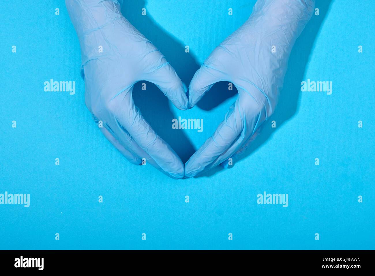 Las manos en guantes de látex de caucho yacen en la forma de un signo de corazón sobre un fondo azul con sombras claras Foto de stock