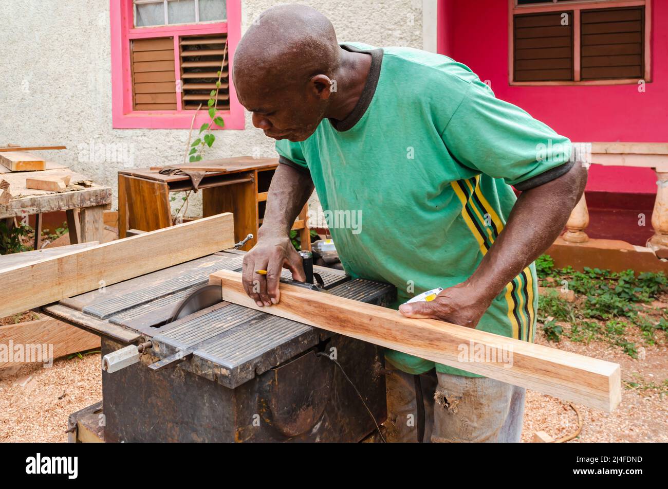 Un cabinate maker con un pencel entre sus dedos se dobla sobre la mesa de  una vieja sierra eléctrica mientras sostiene un pedazo de madera de 2 x 2  que él usa