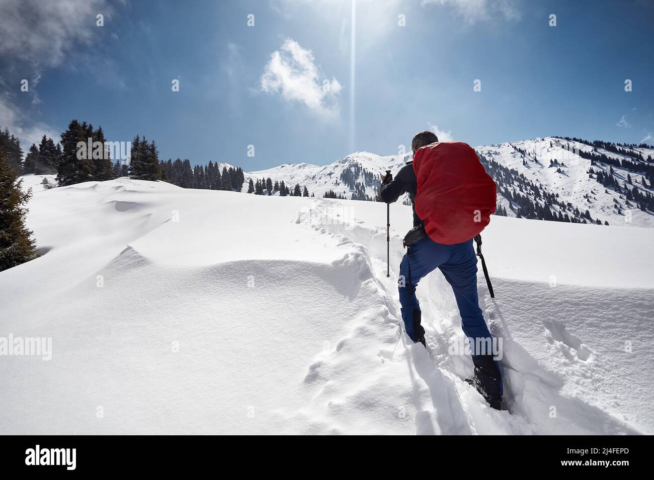 Hombre con mochila caminando en la colina de nieve en las hermosas montañas contra el cielo azul en Almaty, Kazajstán Foto de stock