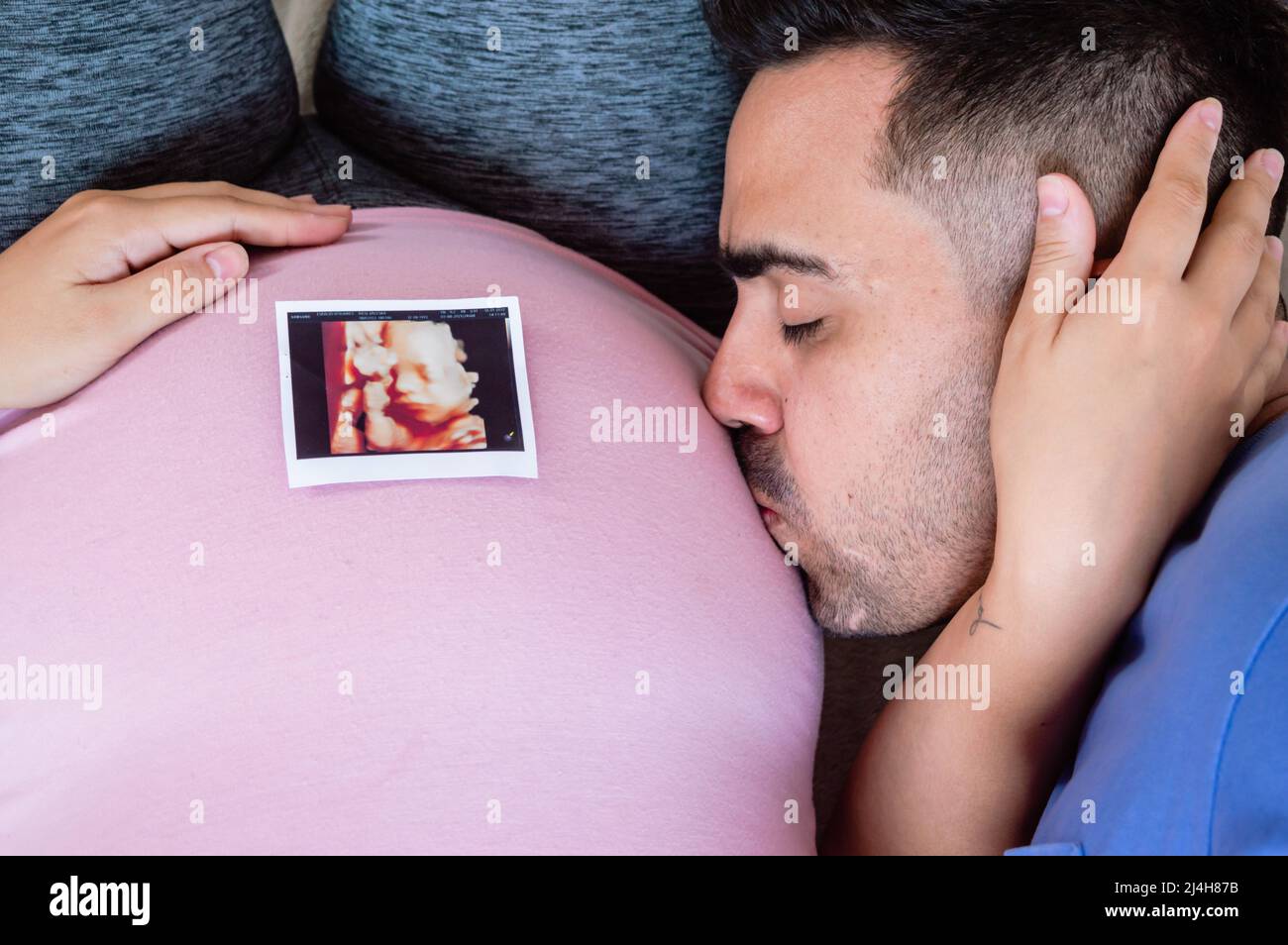 hombre de cerca tumbado en la cama besando el vientre de su esposa embarazada, con la foto de ultrasonido impresa en el vientre. Foto de stock