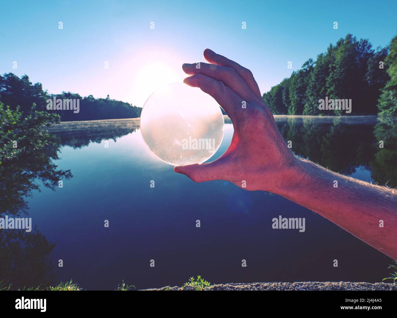 Mirando a través de la lente de cristal. La bola de cristal de sujeción a mano refleja de forma exclusiva la escena del lago durante el verano con la puesta de sol. Foto de stock