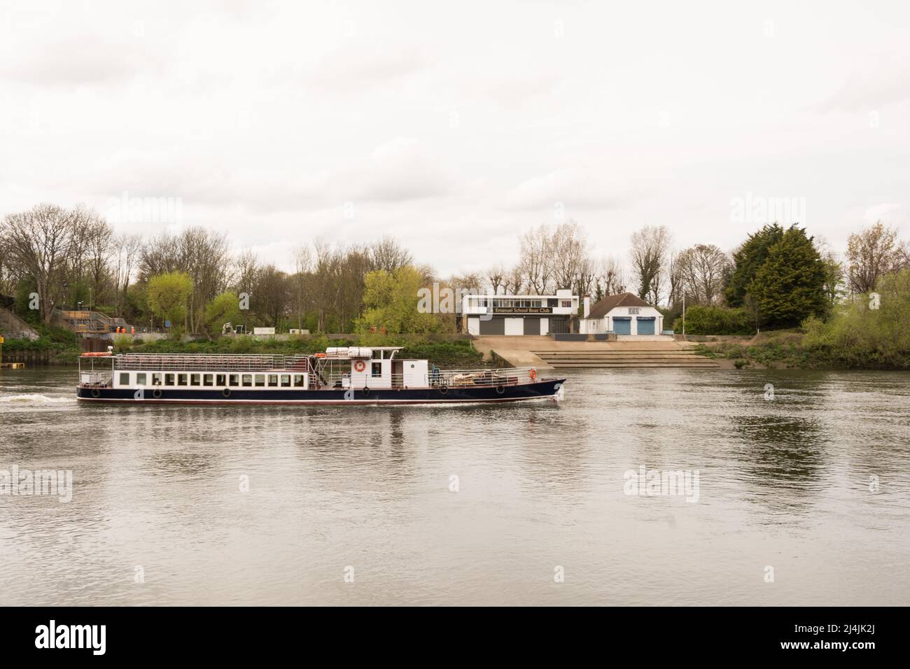 Un crucero de placer vacío pasando frente al Emanuel School Boat Club en un plácido río Támesis en Barnes, al suroeste de Londres, Inglaterra, Reino Unido Foto de stock