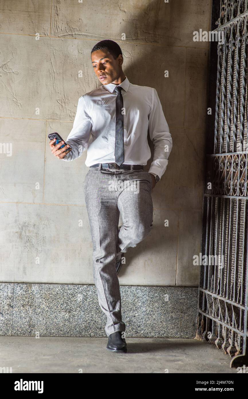 Usando una camisa blanca, una corbata negra, pantalones grises, zapatos de  cuero, apoyado contra una pared, un joven hombre de negocios negro guapo  está parado fuera de un metal Fotografía de stock -