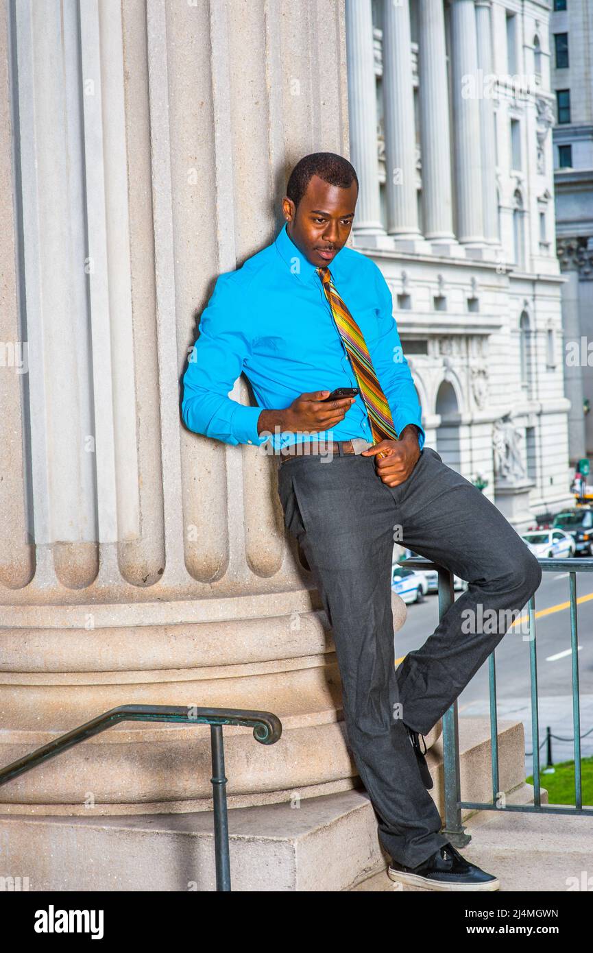 Vestirse en una camisa azul claro, pantalones grises, zapatos de cuero, una  corbata patrón, un joven hombre de negocios negro está parado fuera de un  edificio de oficinas, comprobando Fotografía de stock -