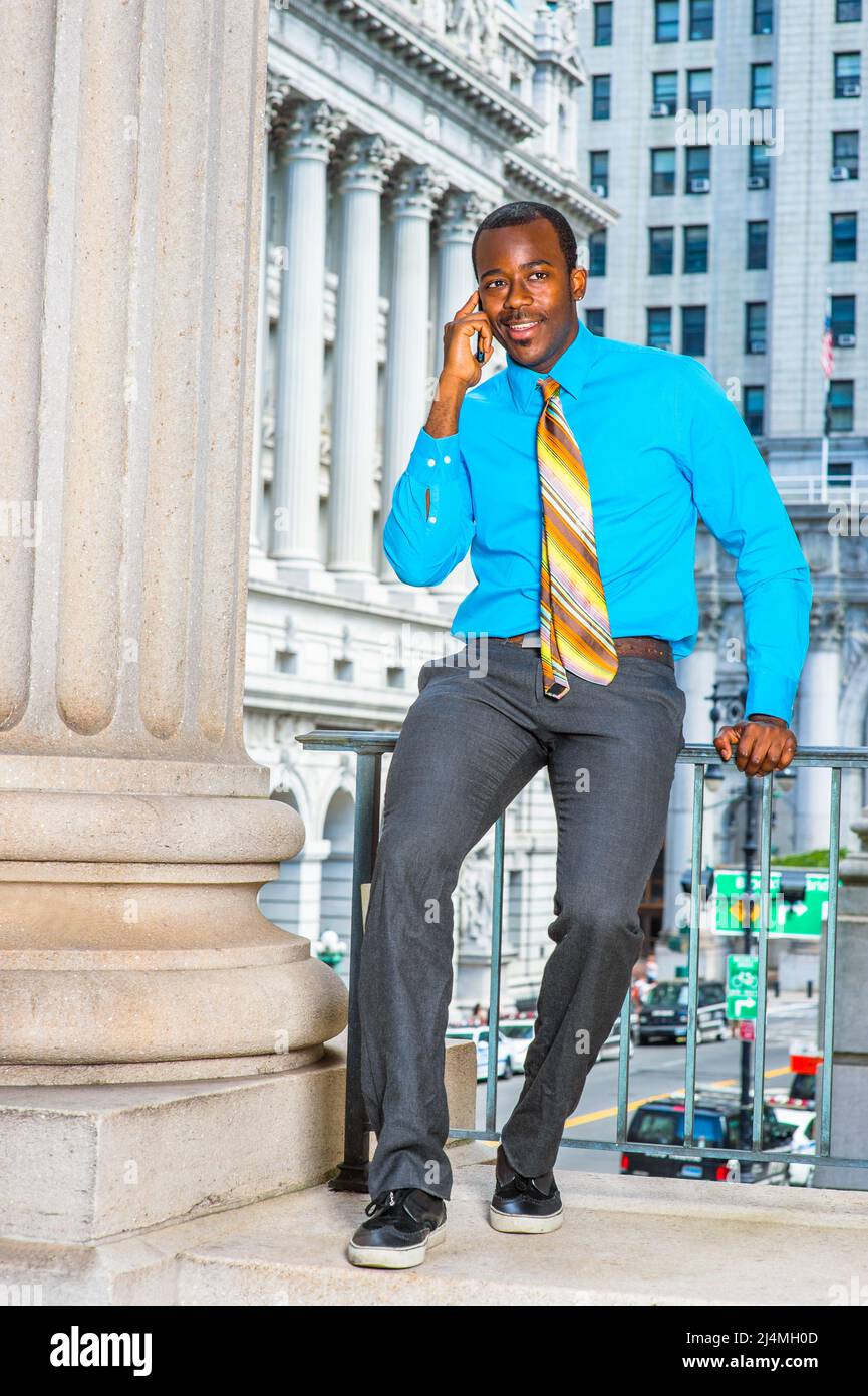 Vestirse en una camisa azul claro, pantalones grises, zapatos de cuero, una  corbata de patrón, un joven hombre de negocios negro está sentado fuera de  un edificio de oficinas, llamando Fotografía de