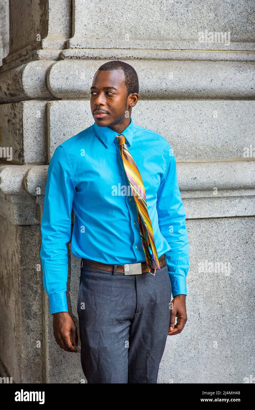 Vestirse en una camisa azul claro, una corbata colorida con patrón,  pantalones grises, zapatos de cuero, un joven hombre de negocios negro está  sentado afuera para tomar un descanso Fotografía de stock -