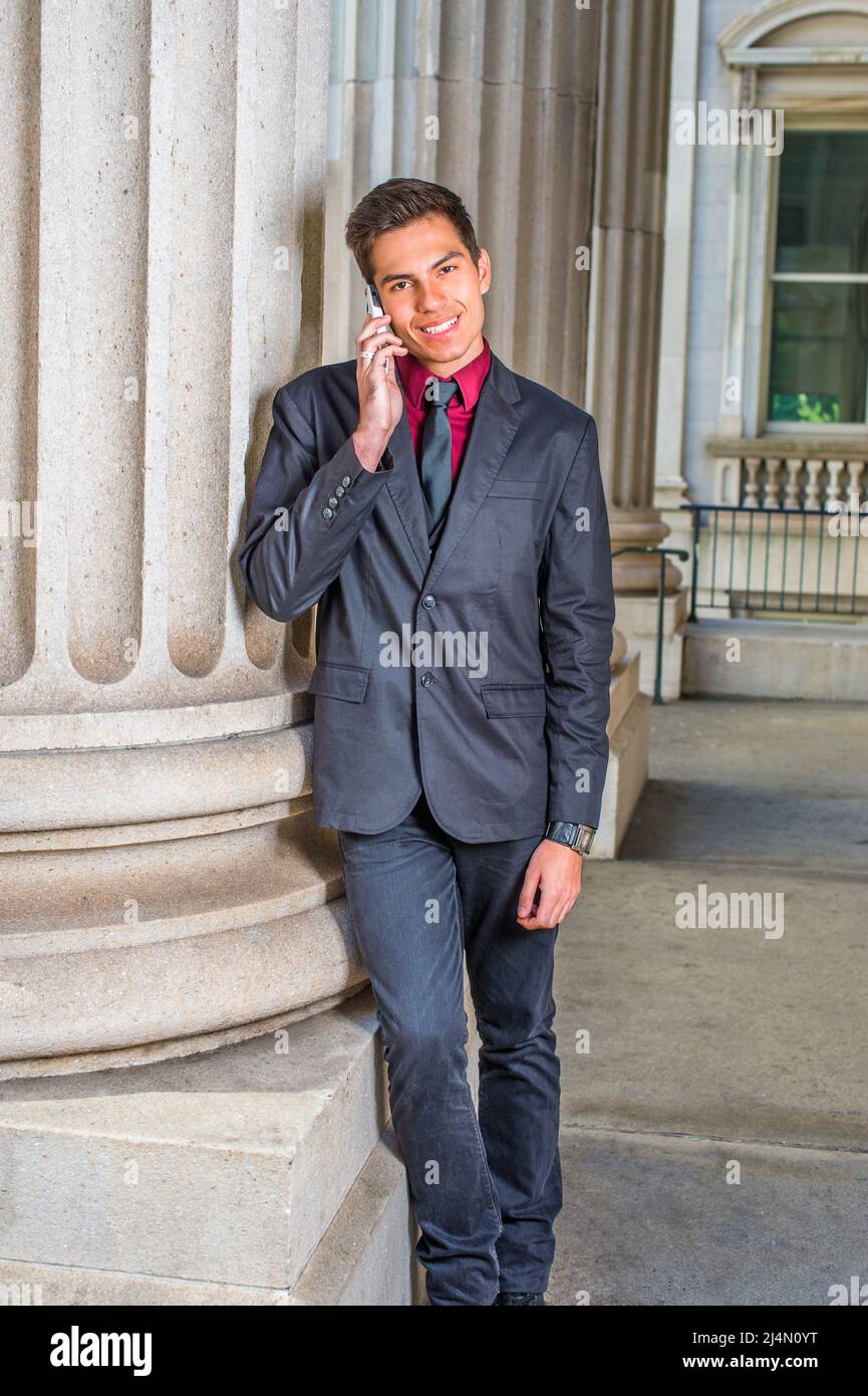Vestirse en una camiseta interior roja, un blazer negro, vaqueros, corbata,  un estudiante joven de la universidad guapo se inclina contra una columna  fuera de una oficina, sonriendo, m Fotografía de stock -