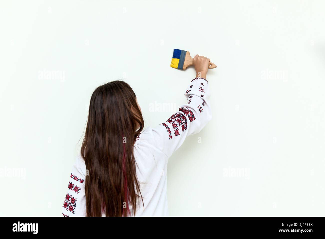 Una chica en un vestido con bordado dibuja en una pared blanca, sostiene un pincel pintado en azul y amarillo. La chica protesta contra la guerra y dibuja la fla Foto de stock