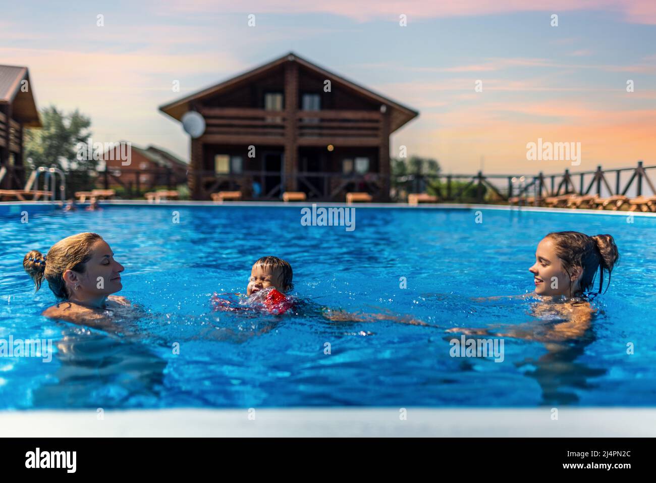 Feliz hermosa alegre mamá juega con sus hijos, hija mayor y pequeño hijo en brillantes mangas en una piscina azul con agua transparente Foto de stock