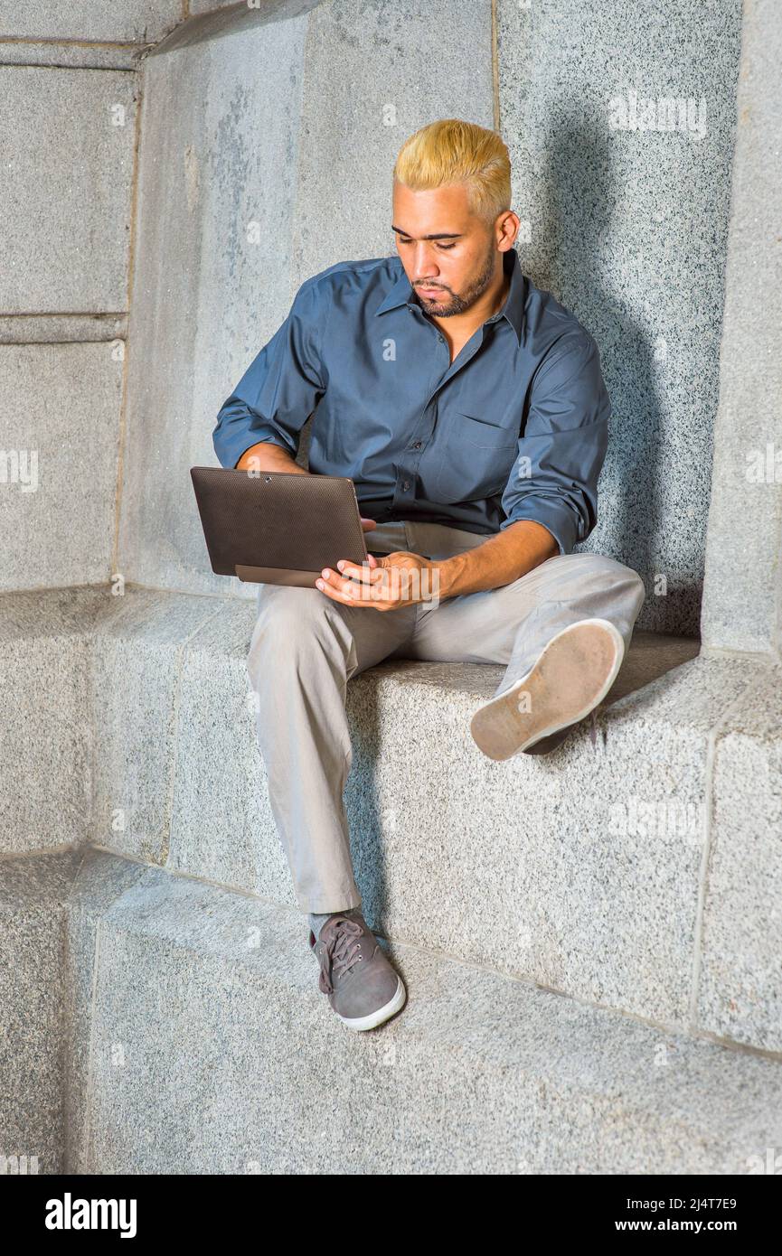Hombre joven trabajando fuera. Usando una camisa azul, pantalones grises,  zapatos informales, un chico joven con barba, el pelo amarillo está sentado  junto a una pared de hormigón, mirando Fotografía de stock -