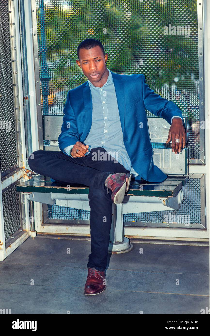 Hombre de negocios infeliz. Usando un blazer azul, pantalones negros,  zapatos de cuero marrón, corte de pelo corto, un individuo negro joven está  sentado en una silla fuera, crossín Fotografía de stock -