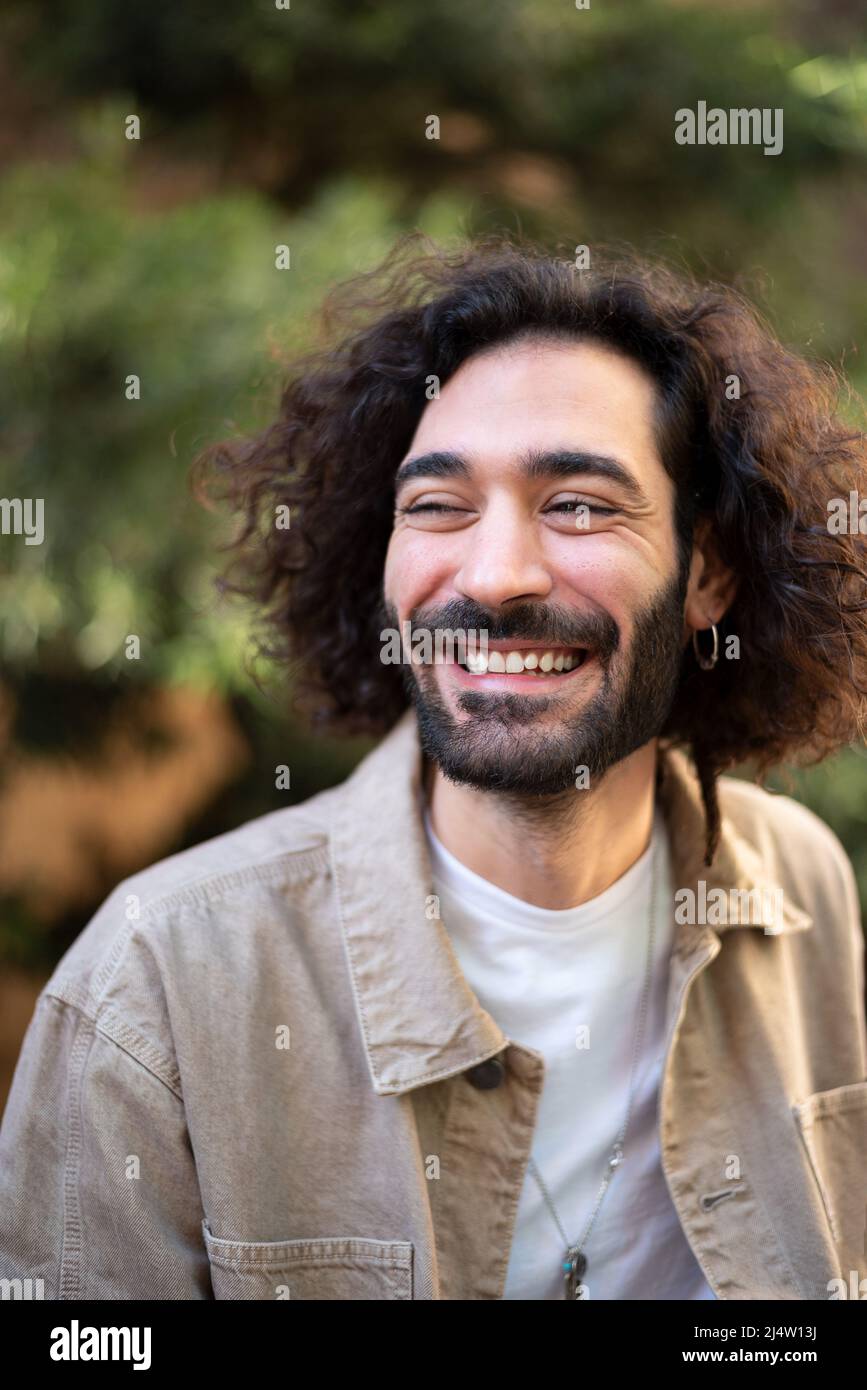 Hombre hispano optimista en ropa informal con pelo rizado y barba mirando hacia fuera y sonriendo alegremente el día de verano en el parque Foto de stock