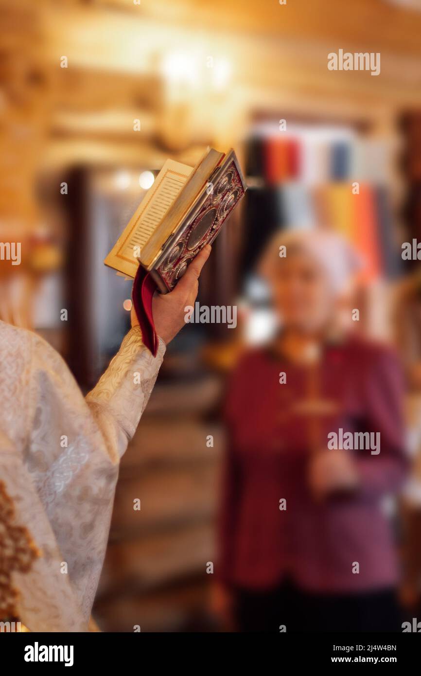 Foto recortada del sacerdote sosteniendo un libro de oración decorado, leyendo Orar durante la ceremonia delante de la mujer sosteniendo la cruz. Foto de stock