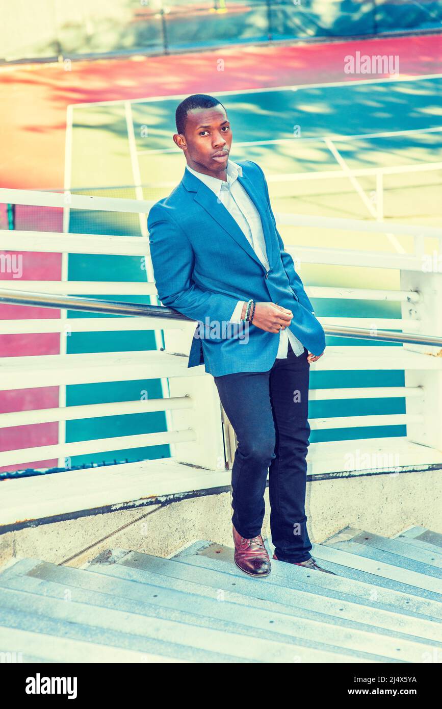 Hombre esperando por ti. Usando un blazer azul, pantalones negros, zapatos  de cuero marrón, corte de pelo corto, un individuo negro joven parado  contra vallas de metal por un tenn Fotografía de