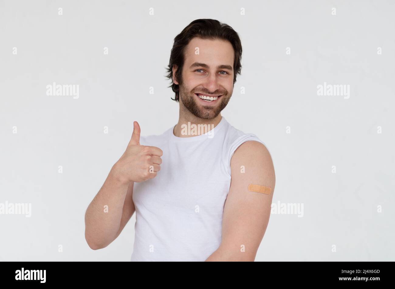 Feliz joven europeo con barba de dos días en la camiseta muestra el hombro después de la vacunación y el pulgar hacia arriba Foto de stock