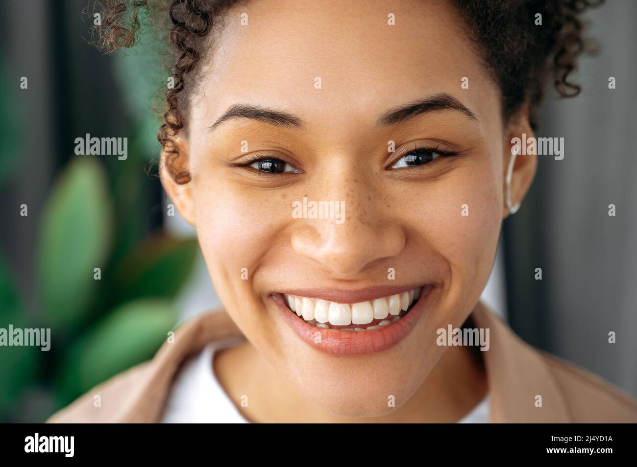 Foto de primer plano de una hermosa mujer joven afroamericana de pelo rizado positivo, chica de raza mixta con pecas, mirando la cámara, sonriendo, mostrando su sonrisa blanca como la nieve Foto de stock