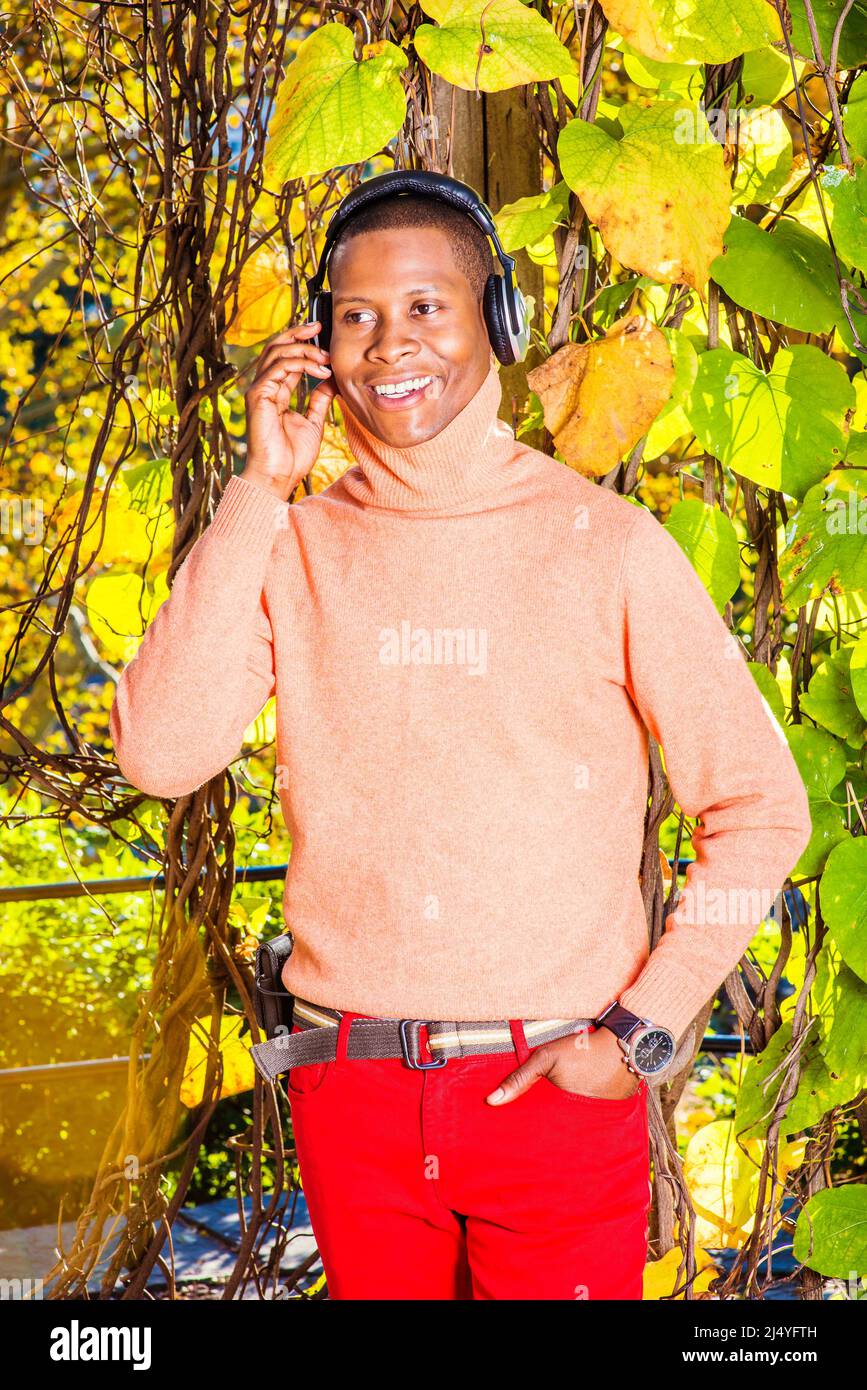 Hombre escuchando música. Vestirse en jersey naranja claro con cuello alto,  pantalones rojos, llevar auriculares, reloj de pulsera, un joven negro está  de pie por b Fotografía de stock - Alamy