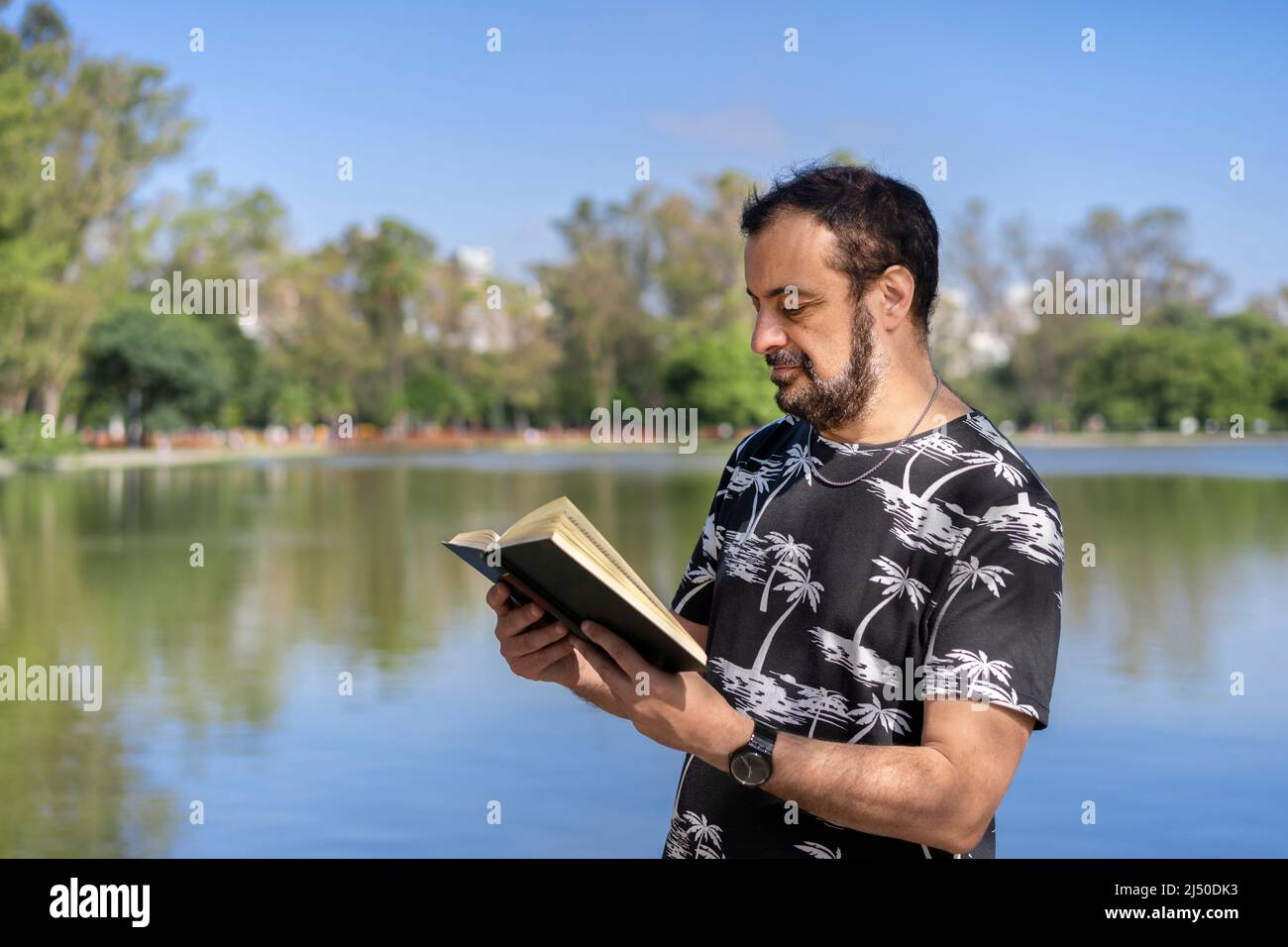 Hombre maduro leyendo junto a un lago, disfrutando de tiempo libre en un día soleado Foto de stock