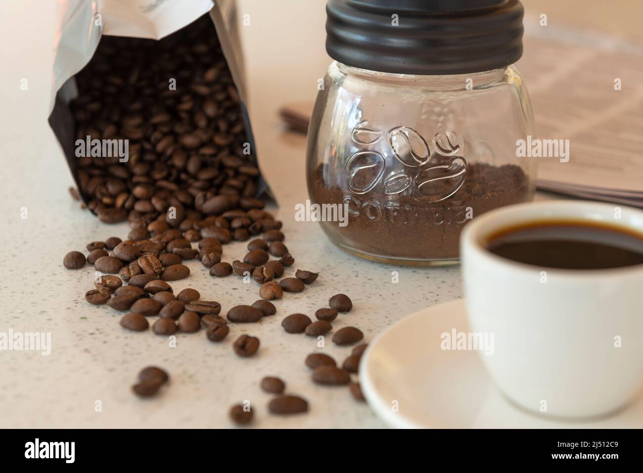 Los granos de café se derraman de su paquete en una mesa de trabajo con una taza de café y café molido cerca. Foto de stock