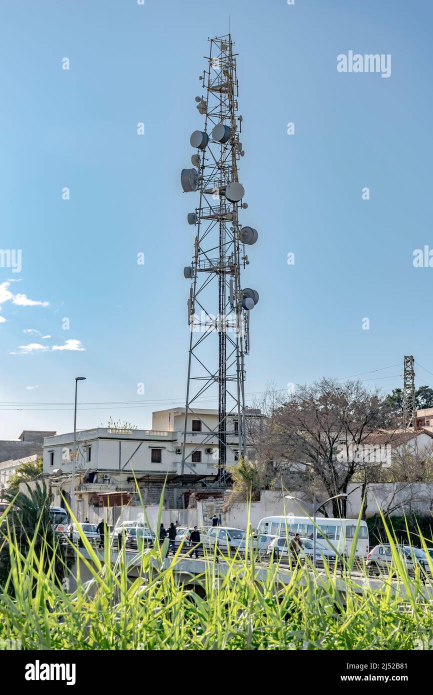 torre de antena de telecomunicaciones en el barrio de una casa. Césped verde en primer plano, personas peatonales, autobuses y coches en el puente de Saoula. Foto de stock