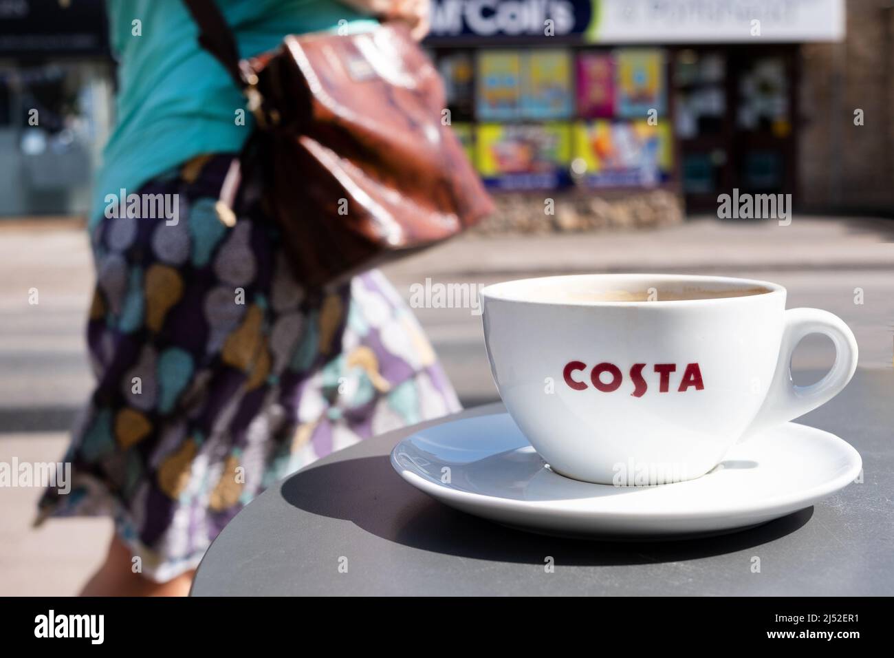 Una taza de café Costa, mostrando claramente el logotipo de la compañía, en una mesa exterior en una calle alta del Reino Unido. Una mujer está pasando detrás de la mesa Foto de stock